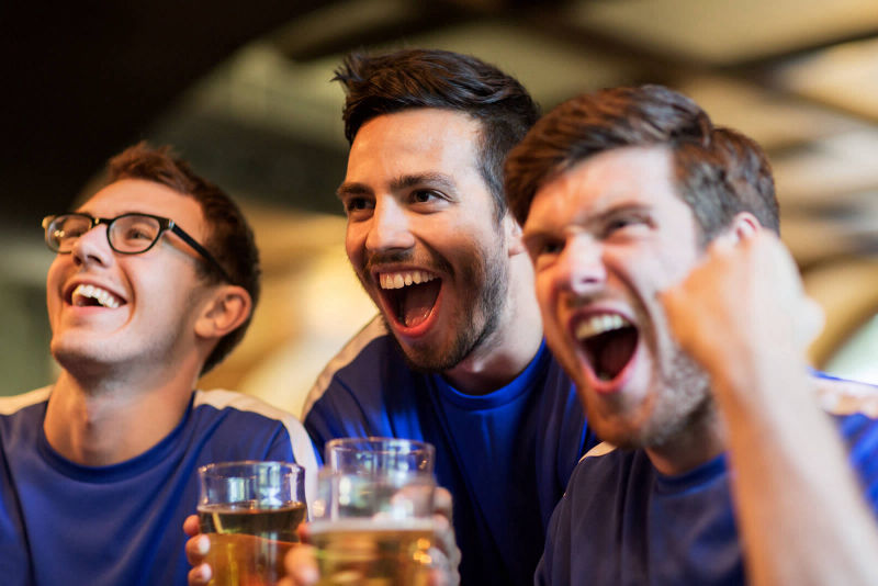 Three guys drinking draft, smiling watching a sports game with excitement.